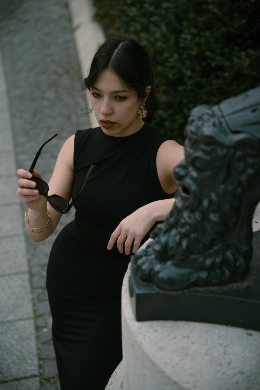 A woman in a black dress holding sunglasses, leaning on a statue