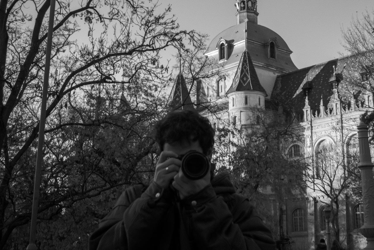 Miraç holding his camera up to his face in front of a castle in Budapest, in black and white