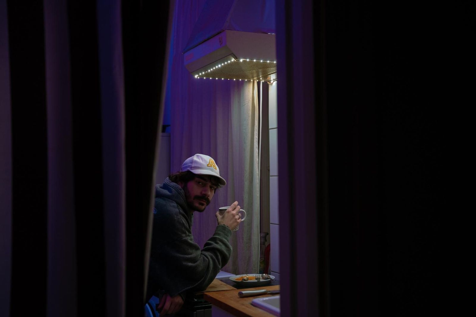 Miraç in a kitchen lit purple, holding a mug, seen between two curtains