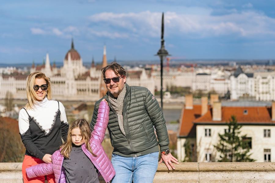 A family with a young girl in a pink jacket above the city, the Parliament behind them
