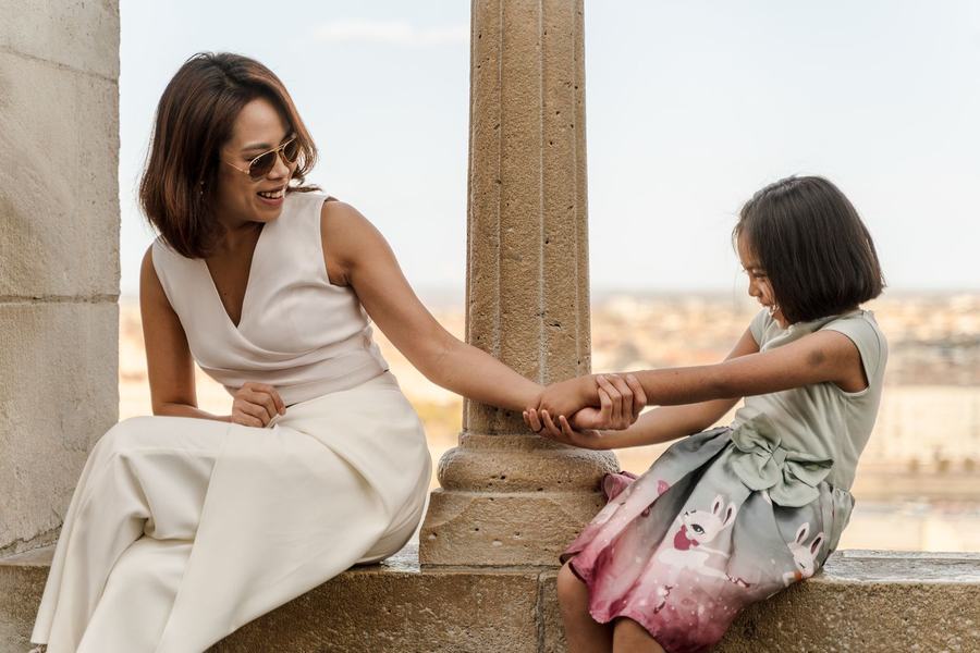 A mother and daughter holding hands across a stone column above the city