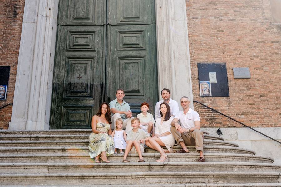 A family of nine sitting on the stone steps of a church with tall green doors in Venice