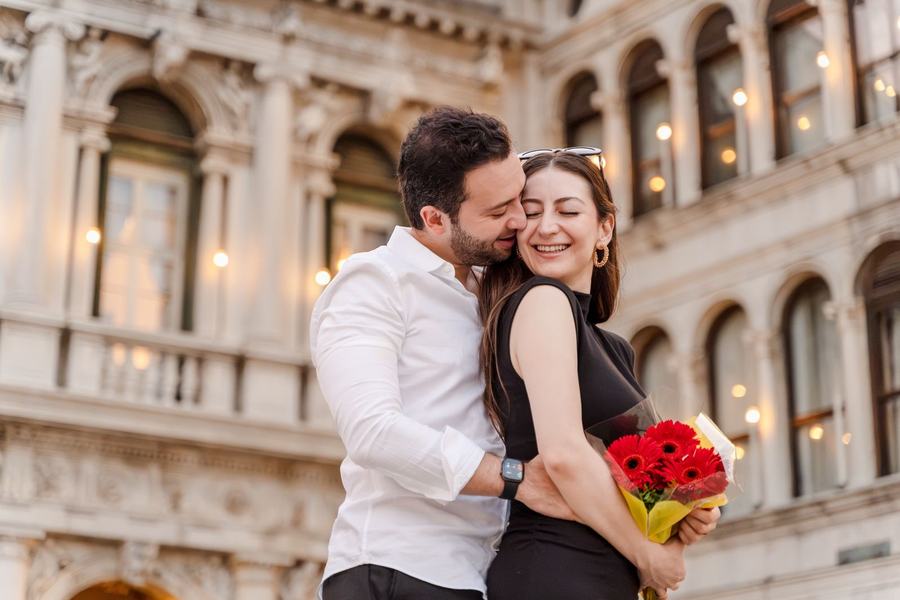 A man hugging a laughing woman holding red flowers in St Mark's Square at dusk