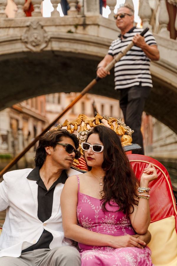 A couple in sunglasses on a gondola below a bridge, the gondolier behind them