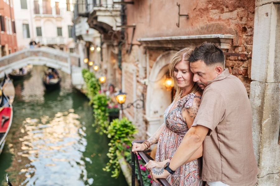 A couple standing close on a small balcony over a canal lit by lanterns