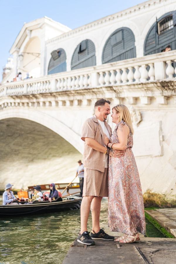 A couple embracing on the bank below the Rialto Bridge as a gondola passes