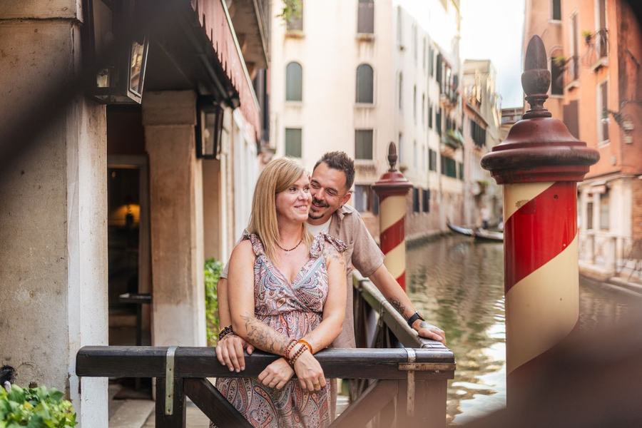 A couple leaning on a wooden railing by a canal with red and white mooring poles