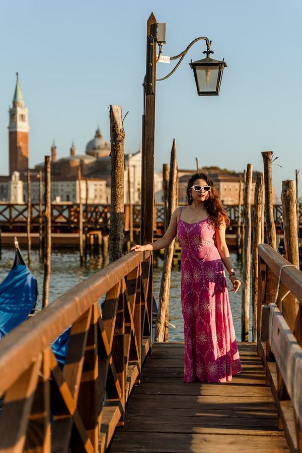 A woman in a pink dress and white sunglasses on a pier beside a lantern, San Giorgio behind her