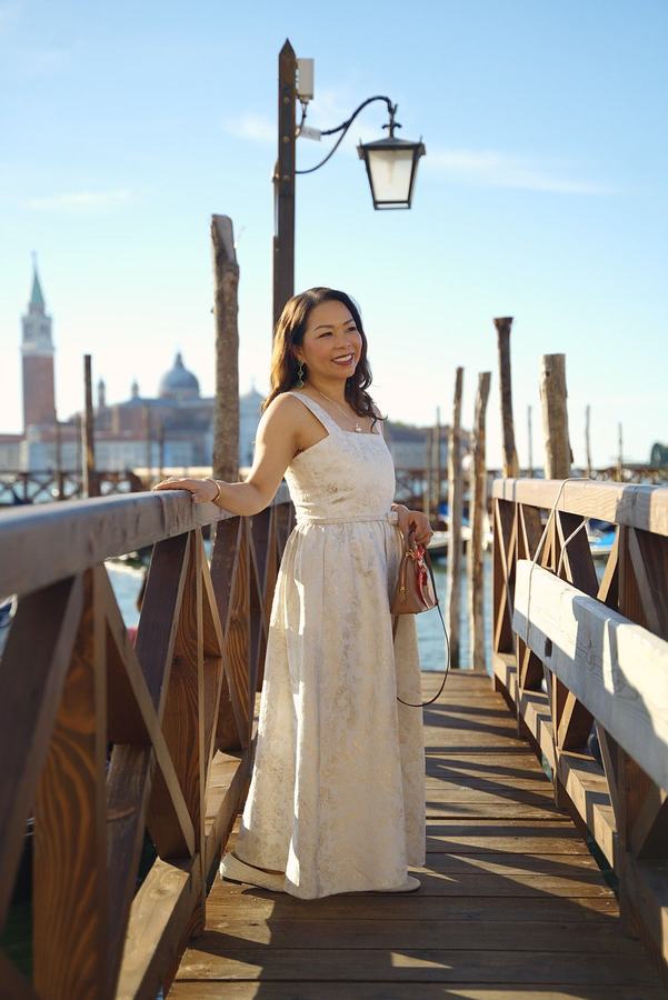A woman in a long white dress on a wooden pier below a lantern, San Giorgio behind her